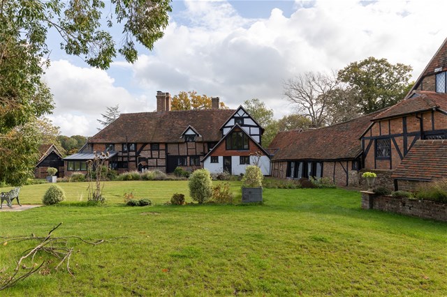 A large traditional English Tudor-style house with black timber framing and white walls, surrounded by a well-kept green lawn, trees, shrubs, and cloudy sky overhead.