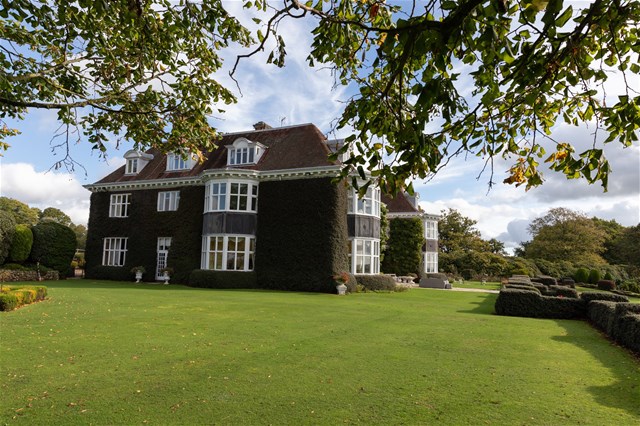 Large ivy-covered mansion with multiple tall windows and gabled roofs, surrounded by well-manicured lawns and neatly trimmed hedges, under a partly cloudy sky and leafy tree branches.