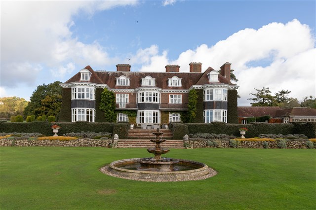A large, elegant mansion with ivy-covered walls, tall windows, and multiple chimneys sits behind a neatly trimmed lawn. A stone fountain is centered in front of the house, surrounded by landscaped gardens.
