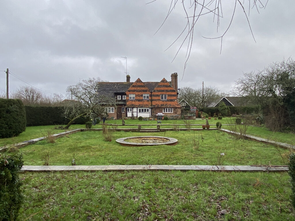A large, red-brick house with prominent chimneys stands at the end of a grassy garden with a circular fountain in the center, surrounded by trees and hedges under a cloudy sky.