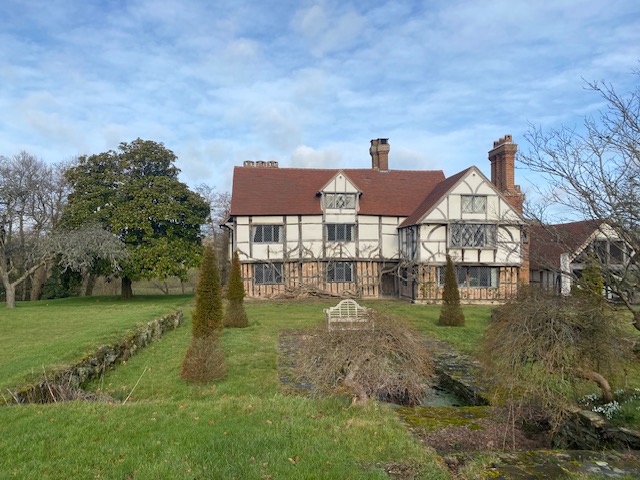 A large Tudor-style house with exposed timber framing and red-tiled roofs sits amid a grassy lawn, with manicured shrubs, leafless trees, and a wooden bench in the foreground under a partly cloudy sky.