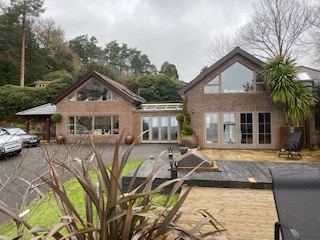 A modern two-story brick house with large windows, surrounded by trees and plants. The front yard features a patio area, a driveway with parked cars, and landscaped greenery under a cloudy sky.