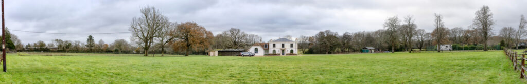 A panoramic view of a grassy field with a white two-story house and a small outbuilding surrounded by leafless trees under a cloudy sky.