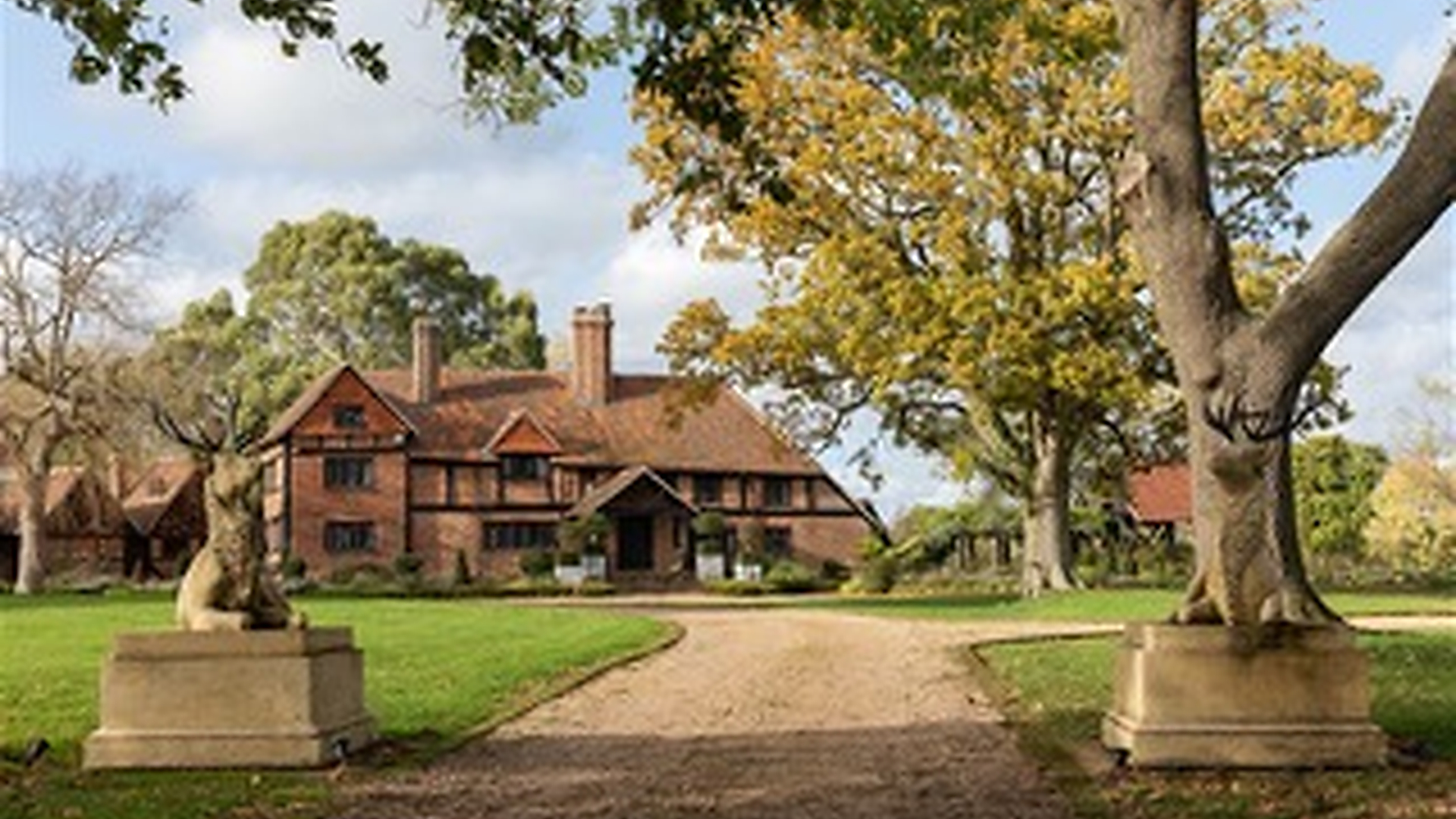 A large brick country house with a red-tiled roof sits at the end of a curving gravel driveway, flanked by two stone deer statues and surrounded by green lawns and tall trees.
