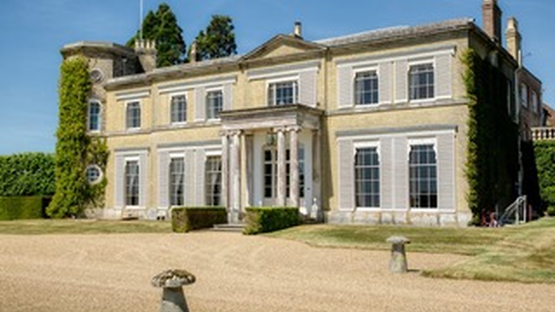 A large, elegant Georgian-style mansion with symmetrical windows, tall white columns at the entrance, light brick walls, and green ivy growing on one side, set on a gravel driveway and manicured lawn.