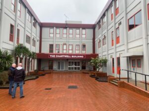 Two people stand talking in a red-bricked courtyard outside a three-story building with red-trimmed windows and a sign reading The Chartwell Building above the entrance. The sky is overcast.