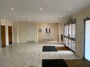 A clean, empty building lobby with tiled floor, white walls, ceiling lights, double entrance doors with glass panels, and brown and black floor mats near the doors.