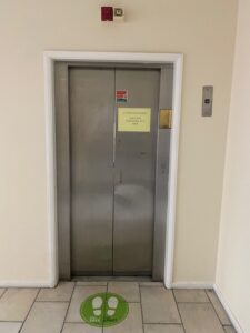 A closed silver elevator door in a beige hallway, with a yellow notice taped to the door and a green floor sticker reading STAY APART showing two footprints in front of the elevator.