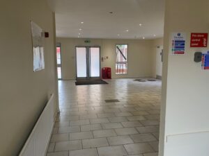 A clean, empty hallway with tiled floors and cream-colored walls leads to a set of glass exit doors. Fire safety signs, a fire alarm, and fire extinguishers are visible near the exit. Natural light enters through the windows.