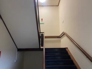 A dimly lit stairwell with dark steps, wooden handrails, and cream walls. There are green emergency exit signs and a red fire alarm box on the upper landing wall.