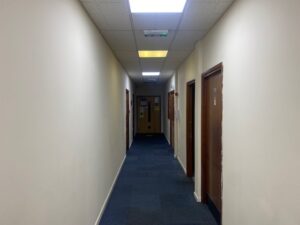 A long, narrow hallway with cream-colored walls, blue carpet, and several wooden doors on both sides, leading to a closed door at the end under fluorescent ceiling lights.