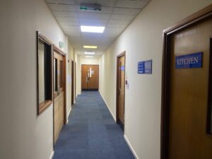 A well-lit hallway with blue carpet and beige walls, featuring several wooden doors, including one labeled KITCHEN and another with an NHS sign. The corridor leads to a closed door at the far end.