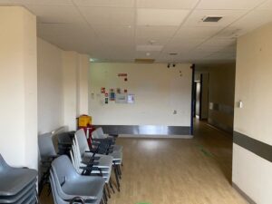 A stack of empty plastic chairs lines the wall in a bare, dimly lit hallway with wood flooring and a drop ceiling. Safety signs and a yellow bin are attached to the far wall. The corridor appears to be in a hospital or clinic.