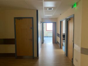 A hallway in a building with wooden doors on both sides, light-colored walls, a wood-patterned floor, and a green exit sign above one doorway; sunlight shines through a window at the end.