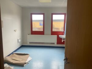 An empty room with blue flooring, two red-framed windows, a white sink on the right wall, a radiator below the windows, and pieces of torn brown paper on the floor. The door is partially open.