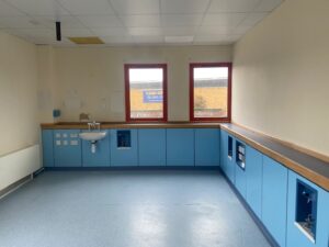 A clean, empty room with blue lower cabinets, a sink on the left wall, two red-framed windows, and white walls and ceiling; outside the windows, part of a building and a sign are visible.