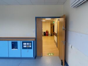 A mostly empty room with blue cabinets and wall outlets, an open wooden door leading to a hallway with light wood floors, bright lighting, and cleaning equipment visible in the distance.