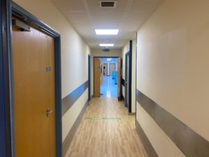 A well-lit hospital corridor with wooden doors on both sides, beige walls, a wood-patterned floor, and a person visible at the far end near a brightly lit room.