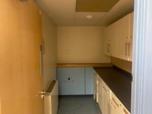 A small, empty room with a countertop, cabinets above and below, a door partially open, and fluorescent ceiling lights. The room appears unused and has minimal decoration.