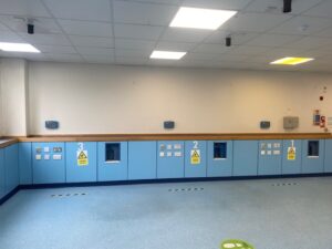 A hospital room with light blue cabinets labeled 1, 2, and 3, various electrical outlets, warning signs, and social distancing floor markings under a white ceiling with fluorescent lights.
