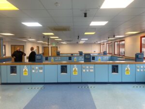 Two men stand and talk in a large, bright room with blue workstations numbered 4, 5, and 6. The workstations have electrical outlets and warning signs. The room has ceiling lights and large windows along one wall.
