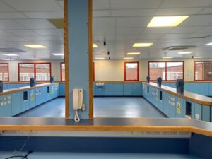 A bright, empty laboratory with blue workstations, red-framed windows, ceiling lights, a white wall phone, and a central sink. Electrical outlets and safety signs are visible along the counters.