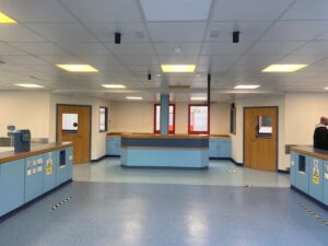 A clean, empty hospital or clinic reception area with blue countertops, wooden doors, light blue flooring, and red window frames. One person stands near the right door; overhead lights illuminate the space.