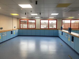 A spacious, empty room with blue flooring and cabinets, bright overhead lighting, red-framed windows, and caution signs on some cabinets. The windows overlook buildings outside.