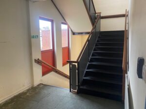 A stairwell with black steps and wooden handrails, featuring two large windows with red panels and a green emergency exit sign on the wall. Light streams in through the windows, illuminating the area.