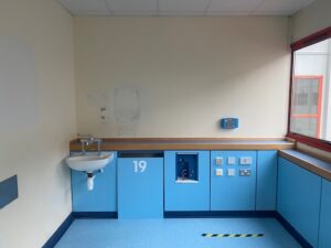 A hospital room corner with blue cabinets, a small white sink, electrical outlets, a window to the right, and the number 19 on a cabinet door. The floor and cabinets are blue, and yellow-black tape marks the floor.