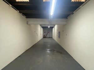 A long, empty corridor with white walls, a gray floor, and a dark exposed ceiling. Fluorescent lights hang overhead, and a red ladder is visible at the far end near the back wall.