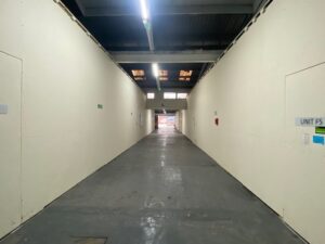 A long, empty corridor with plain white walls and a gray floor, lit by overhead lights. Signs and a fire extinguisher are mounted on the walls, and daylight is visible at the far end.