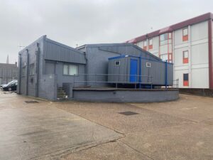 A grey industrial building with a blue portable cabin attached, set on a concrete lot. A red and white apartment building is visible in the background under a cloudy sky.