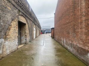 A wet, empty alleyway between a brick archway on the left and a tall brick wall on the right, leading to a distant industrial building under a cloudy, gray sky.