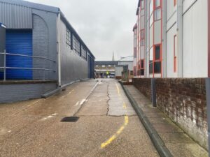 A narrow, wet alleyway between gray industrial and white buildings leads to shops in the distance, with a church spire visible under a cloudy sky. Yellow lines mark the path along the rough, paved ground.