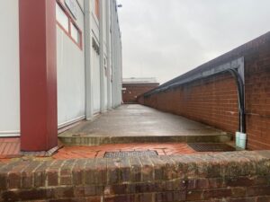 A wet concrete walkway runs alongside a brick wall and a building with white and red panels on a cloudy day. Exposed pipes and cables are visible on the right side of the wall.