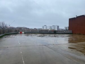 An empty, wet parking lot on a cloudy day with city buildings and a crane in the background. The ground is reflective from rain, and there is a wooden fence along the perimeter.