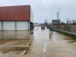 Two people in safety vests walk near a brown car on a wet, empty industrial lot with warehouses, city buildings, and a crane visible in the background under an overcast sky.