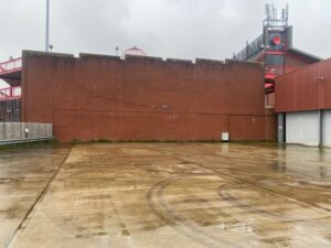 A wet, empty concrete lot with faint tire marks in front of a tall brick wall. Red metal structures and a gray, overcast sky are visible in the background.