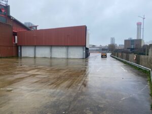 A wet, empty concrete lot beside a large, red industrial building on a cloudy day. A single van drives away toward city buildings and cranes in the background.