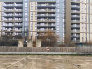 A modern apartment building with balconies stands behind a wooden fence, some trees, and a wet concrete surface. A life ring hangs on the fence in the center of the image.