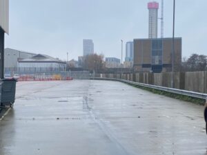 A wet, empty concrete lot with a metal fence, bins on the left, and modern buildings and a tall red-and-white tower in the background on a cloudy day.