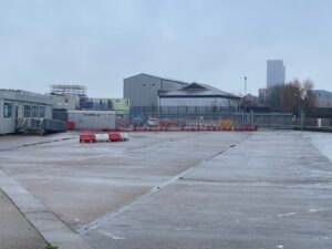 A grey, rainy day at an empty industrial lot with wet pavement, red barriers, and metal fencing. Industrial buildings and a tall modern structure are visible in the background.