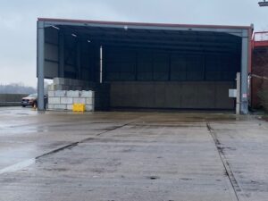 A large, open-sided industrial shed with stacks of concrete blocks inside, a yellow container, and a parked car partially visible on the left. The ground is wet, and the sky appears overcast.
