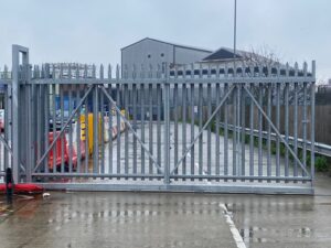 A tall, silver metal security gate blocks a paved road on a cloudy day, with industrial buildings and fencing visible in the background.